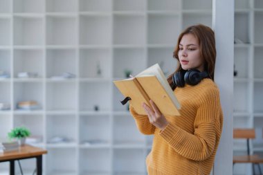 Kulaklıklı mutlu genç kadın bir kitaba dalmış, mutlu bir şekilde öğreniyor..