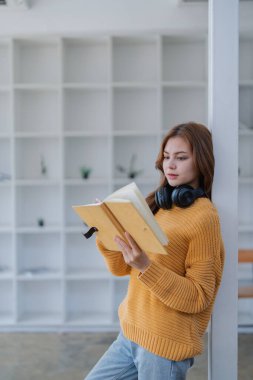 Kulaklıklı mutlu genç kadın bir kitaba dalmış, mutlu bir şekilde öğreniyor..