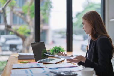 Businesswoman reviews paperwork and laptop on cluttered office desk now..