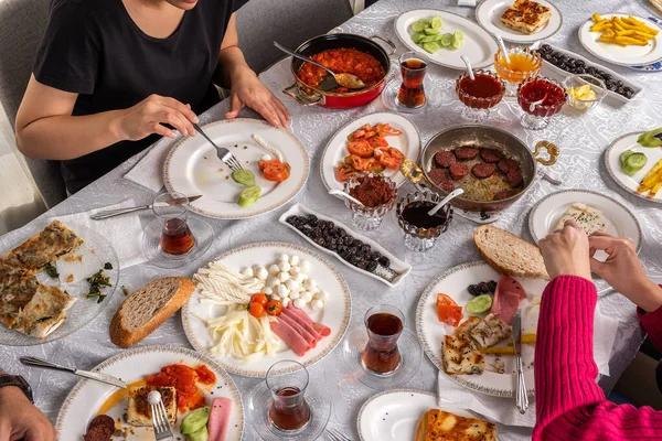 Yummy full breakfast table. Family or friends having brunch. Traditional Turkish breakfast. Drinking tea eating sausage called sucuk. Different cheese, black olive, tomatoes, bread, su borek, pastry.