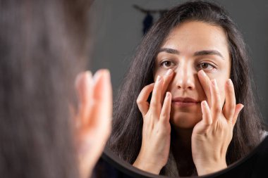 Touching face, millennial brunette woman touching face. Facial skin care, everyday spa treatment concept idea image, enjoy soft moisturized hydrated skin. Over shoulder view girl looking to mirror.