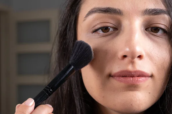 Applying face powder, caucasian brunette woman looking to camera and applying face powder. Close up portrait of girl holding make up brush, dry cosmetic tonal foundation on her face concept idea.