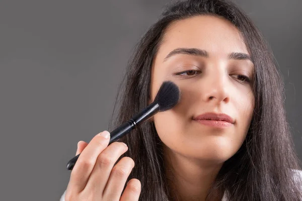 Applying blush, young caucasian girl applying blush. Face powders, using brush for beige foundation to cheekbones. Isolated on dark gray background. Feeling beautiful, perfect makeup concept idea.