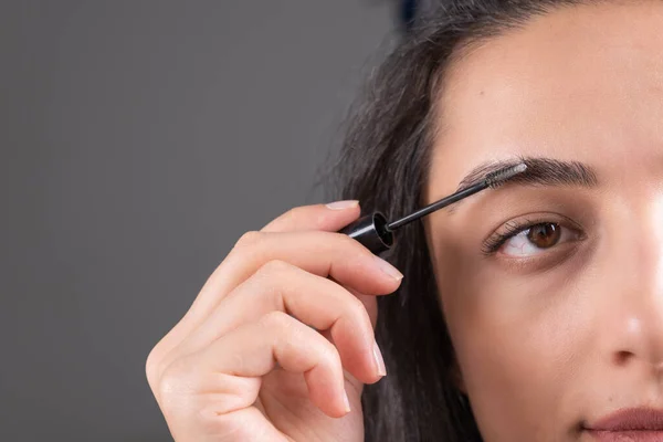 Shaping eyebrow, close up image of brown haired brunette woman shaping eyebrow. Using mascara. Woman making her own make up. Cropped photo, girl getting ready for evening. Dark gray background.