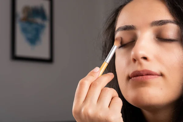 Applying eye shadow, close up image of beautiful woman applying eye shadow. Caucasian brown hair girl making her own make up. Painting cosmetics of young beauty model. Holding brush. Fashion concept.