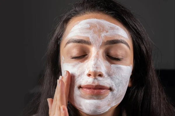Applying clay mask, calmly closed up eyes while applying clay mask. Caucasian girl making spa at home, Beauty treatments concept idea image. Touching her cheek gently. Cosmetic procedures, skin care.