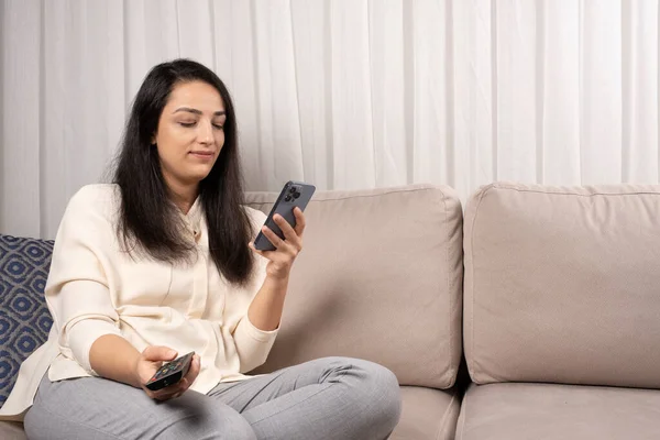 Phone or social media addiction, portrait of caucasian woman sitting on the couch holding tv remote looking smartphone. Internet, Phone or social media addiction concept idea photo, copy space.