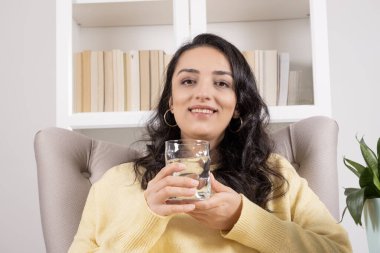 Holding glass of water, happy smiling woman holding glass of water. Head shot portrait of caucasian brunette recommending pure mineral fresh water. Sitting on armchair at home. Healthy lifestyle.