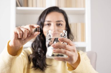 Capsule of vitamin, head shot portrait of caucasian woman showing capsule of vitamin. Selective focus on hands. Holding glass of water, smiling happy millennial girl. Health care concept idea image.