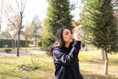 Workout in autumn in park, woman runner rest after workout in autumn in park. Young attractive brunette lady drinking water from glass bottle. Walking, refreshing against green nature background.