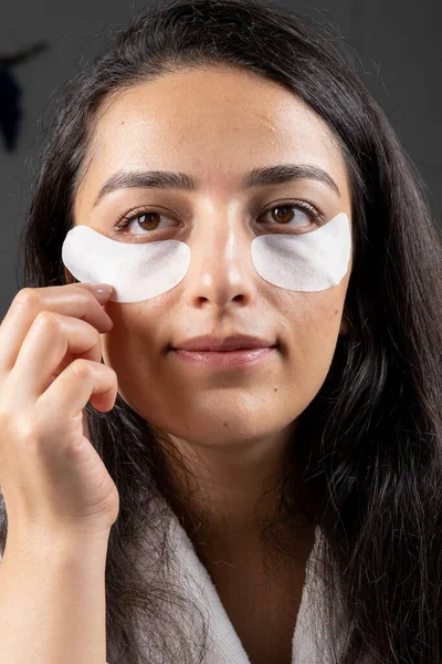 Under eye patches, close up caucasian millennial brunette woman applying under eye patches. Looking mirror using anti aging cosmetic skin care beauty routine at home. Daily under eye bag treatment.