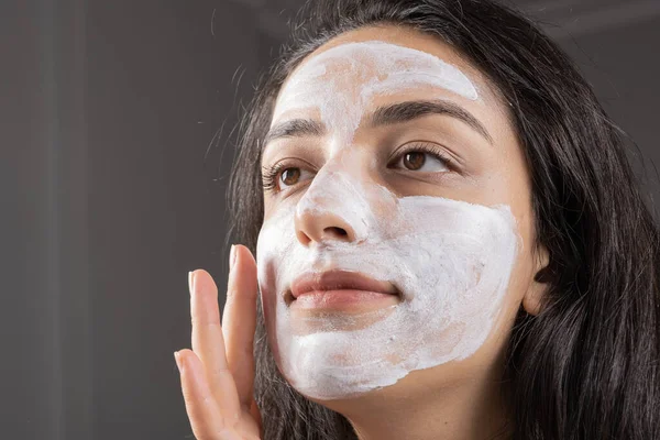 Clay beauty mask, portrait of young brunette woman applying clay beauty mask. Spa beauty treatments at home. Copy space. Beautiful millennial girl with cosmetics on her face. Skin care concept idea.