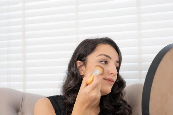 Applying blush, brunette millennial woman applying blush. Beige foundation powder on cheeks. Looking mirror holding, using make up brush. Sitting armchair at home. Putting daily make up concept idea.