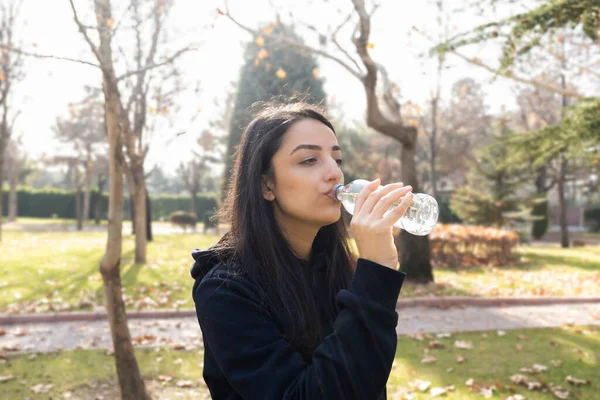 Drinking water, profile view of sporty woman drinking water. Holding glass bottle. Walking or jogging in park. Sunny autumn day. Copy space. Fit girl enjoying aqua. Healthy lifestyle concept idea.