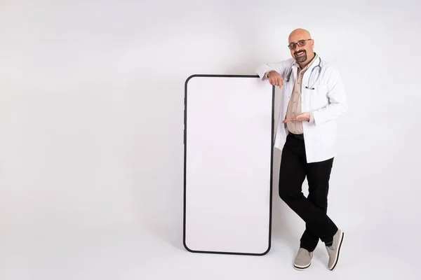 Medical app concept idea, caucasian male doctor leaning at big smartphone with blank screen for mock up. Isolated on light gray background. Male doctor demonstrating copy space for advertisement.