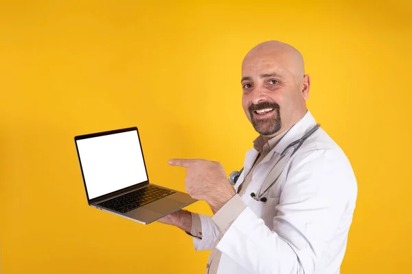 Doctor showing empty white screen of modern laptop for mock up. Online appointment or consultation concept idea. Isolated orange background. Male medical doctor in uniform points finger at display.