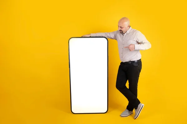 Big blank smartphone, full body length of man pointing finger at big blank smartphone. Human sized mobile phone mock up. Yellow background. Smiling guy showing empty white screen. Ad concept idea.