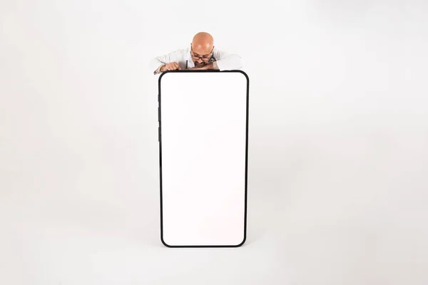 Big smartphone mock up, caucasian man leaning big smartphone mock up from behind. Light gray background. Copy space. Guy looking  from top white blank screen of life size cell phone.
