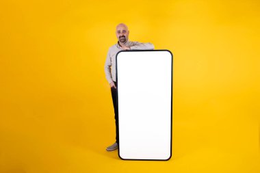 Tavsiye edilen konsept fikir, beyaz tenli yakışıklı erkek büyük akıllı telefona eğilip konsept fikir öneriyor. Beyaz ekranlı büyük bir cep telefonu. Sarı stüdyo arka planda bekliyor. Taşınabilir teklif.