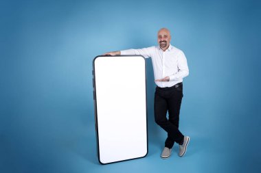 Mobil uygulama reklam konsepti, mutlu adam tüm vücut boyu büyük cep telefonuna yaslanıyor. Modern akıllı telefon fikri uydurma. Mavi stüdyo arka planında duruyor. Beyaz boş ekran, ekran. Boşluğu kopyala.
