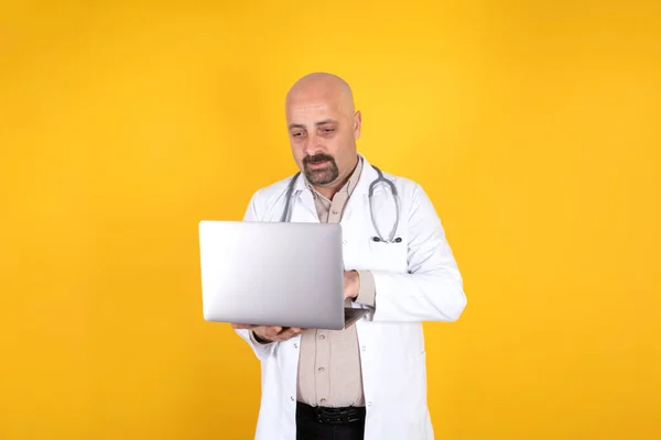 Doctor using laptop, male bald bearded doctor using laptop. Standing isolated yellow background. Holding his notebook computer, typing. Wearing white uniform, coat with stethoscope. Medical concept.