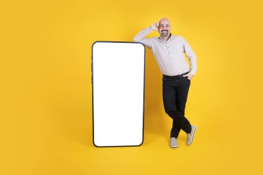 Neşeli bir adam büyük bir cep telefonuna eğiliyor, rahat giyimli, uzun boylu, neşeli bir adam büyük bir cep telefonuna eğiliyor. Beyaz ekran cep telefonu modeli. Smartphone uygulaması, web sitesi önerisi.