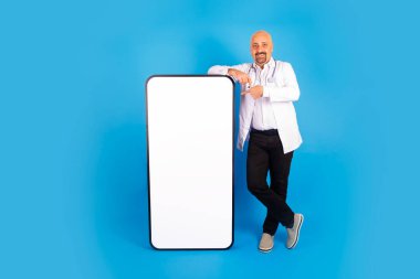Doktor akıllı telefona eğiliyor, beyaz bir doktorun akıllı telefona eğildiği bir vücut boyu fotoğrafı. Mavi stüdyo arka planı, kopyalama alanı. Gülümseyen hastane, tıp hekimi tıp başvurusu.