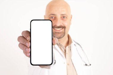 Reklam fikri, doktorun boş ekranlı cep telefonu göstermesi. Olumlu, gülümseyen, mutlu doktor elinde modern akıllı telefon tutuyor. İzole edilmiş beyaz arka plan. Uzayı kopyala steteskop takıyor..
