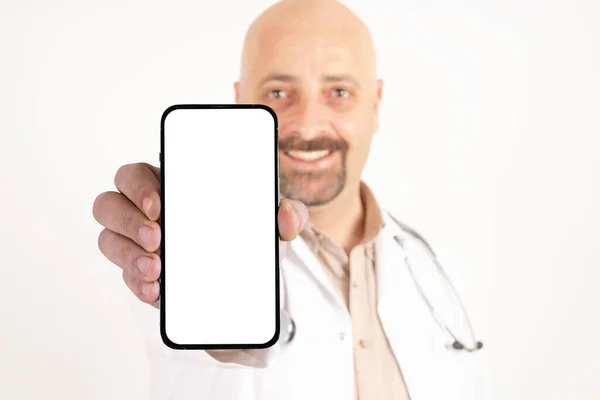 広告コンセプトのアイデア 医師は空の空白の画面の携帯電話を示しています ポジティブで笑顔で幸せな医師が現代のスマートフォンを持っています 隔離された白い背景 コピースペース 聴診器をつけて — ストック写真
