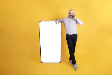 Orta yaşlı beyaz bir adam model almak için boş beyaz ekran ile büyük bir akıllı telefona yaslanıyor. Telefonda konuşuyordu. Sarı arka planda duran küçük göbekli adam, boşluğu kopyala.