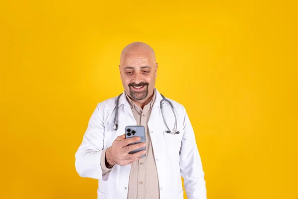 General practitioner doctor holding telephone in his hand. Middle aged guy using technology. Positive smiling physician messaging. Using health clinic application. Yellow background, copy space.