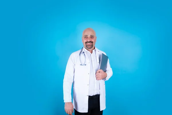 Middle aged doctor in white robe, caucasian bald standing middle aged doctor in white robe. Looking and smiling camera. Holding laptop next to chest. Isolated on blue background, copy space.