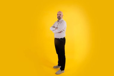 Tam vücut portresi, kel orta yaşlı, gülümseyen insan portresi. İzole edilmiş sarı arkaplan, kopyalama alanı. Sıradan giyinmiş, baba figürü erkek, kolları bağlı. Gururlu, kendinden emin adam..
