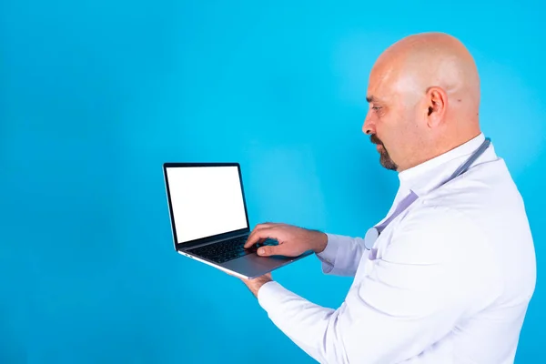 Doctor using laptop, side view portrait of middle aged caucasian doctor using laptop. Blank white screen notebook for mock up. Tele medicine concept idea. Isolated blue background. Online appointment.