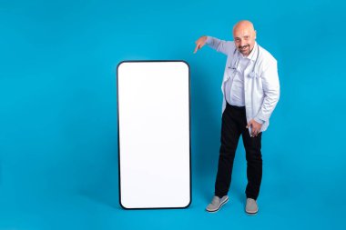 Büyük akıllı telefonun yanında boş ekranlı, tam boy bir doktor duruyor. Hastane web sitesi, tıbbi uygulama, tele tıp fikri. Reklam şablonu. Mavi stüdyo arka planında izole edilmiş. Modern model..
