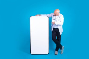 Orta yaşlı erkek doktor, eğiliyor ve kocaman cep telefonunu boş ekrana doğrultuyor. Tam boy bir hekim, izole edilmiş mavi stüdyo arka planında duruyor. Sağlık sigortası fikri, alanı kopyala.