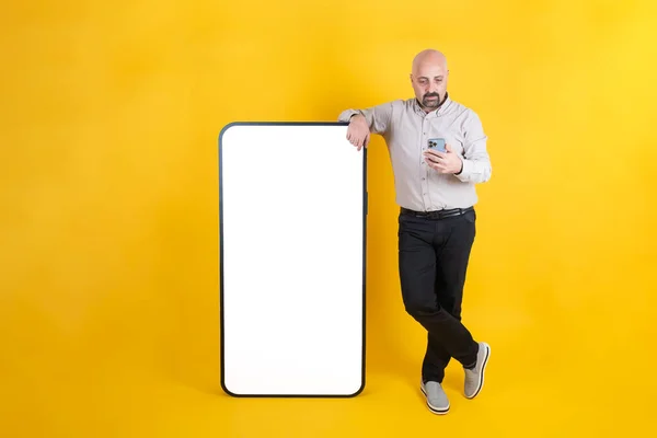 Mobile advertising, middle aged casual dressed man leaning big  huge smartphone for mobile advertising. White blank empty screen mobile phone mock up. Yellow studio background, isolated. Copy space.