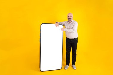 Tüm vücut uzunluğunda, akıllı telefona yaslanmış beyaz ekrandaki panele parmağıyla dokunan bir adam. Kameraya bak. Cep telefonu modeli. Sarı arka plan. Yemek siparişi, afiş.