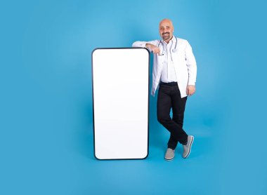 Başvuru ilanı, orta yaşlı doktor başvuru reklamı için akıllı telefona yaslanıyor. İzole edilmiş mavi arka plan. Hastane üniforması ve steteskop giyen bir doktor. Cep telefonu maketi.