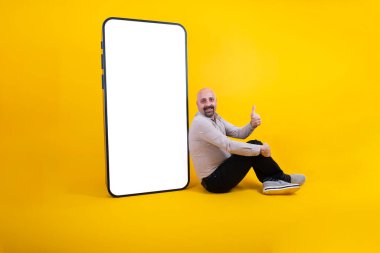 Mobil reklam, orta yaşlı beyaz erkek. Mobil reklam için akıllı telefonun yanında oturuyor. Yeni başvuru formunu sunuyorum. Adam onay konseptini gösteriyor. Sarı arkaplan, boşluğu kopyala.