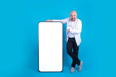 Büyük akıllı telefon, gülümseyen orta yaşlı doktor akıllı telefonu işaret ediyor. Kel hekim boş ekran ile kocaman bir cep telefonuna yaslanıyor. İzole edilmiş mavi stüdyo arkaplanı, kopyalama alanı. 