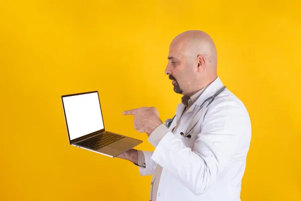 指向笔记本电脑屏幕 中年秃头医生指向笔记本电脑屏幕 白人男性医生拿着带有空白白屏的现代笔记本进行模拟 站在演播室黄色背景之上 — 图库照片