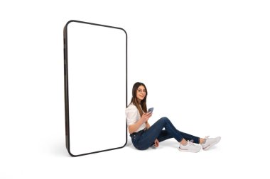 Akıllı telefonlu boş ekran, gülümseyen genç bir kadın yerde oturmuş akıllı telefonlu boş ekran modelini taklit ediyor. Kamera, cep telefonu, internet reklamı, uygulama.