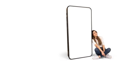 Web sitesi ya da mobil uygulama arayüzü değişimi için boş ekranı olan büyük bir akıllı telefon modeli. Beyaz tenli kadın oturuyor ve cep telefonuna yaslanıyor. Mutlu neşeli güzel kız