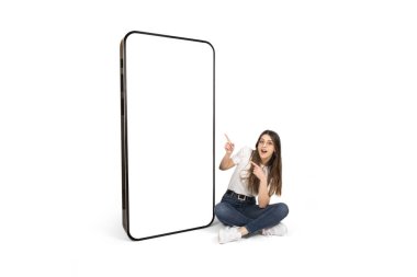 Büyük bir akıllı telefon ekranı, büyük bir akıllı telefon ekranı sergileyen mutlu beyaz kadın. Boş beyaz ekranda büyük bir cep telefonu görüntüsü var. İzole edilmiş beyaz, tam vücut ölçülerinde oturan bir kız..
