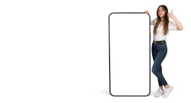 Mobil başvuru ya da web sitesi öneren bir kadının tam vücut fotoğrafı. Beyaz kadın boş ekran modeliyle akıllı telefona yaslanıyor. Baş parmak pozu veriyor. İzole edilmiş beyaz arkaplan, kopyalama alanı. 