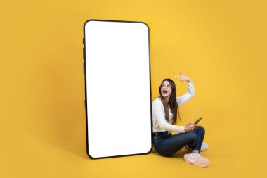Yerde oturan kadın büyük boş ekranlı cep telefonu modelleme alanını işaret ediyor. Akıllı telefon kullanıyoruz. Gülümseyen neşeli kız başvuru öneriyor. Boşluğu kopyala.