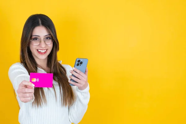 Easy online money transfer,  portrait of caucasian young girl holding smartphone and showing pink debit credit card. Online payment concept idea. Standing over isolated orange studio background.