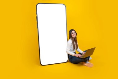 Kocaman bir akıllı telefon modeli, uzun boylu, akıllı telefonlu bir kadın oturuyor. Modern dizüstü bilgisayar, gülümseyen kamera. Sarı stüdyo arka plan, kopyalama alanı. Konsept fikri öneriliyor.