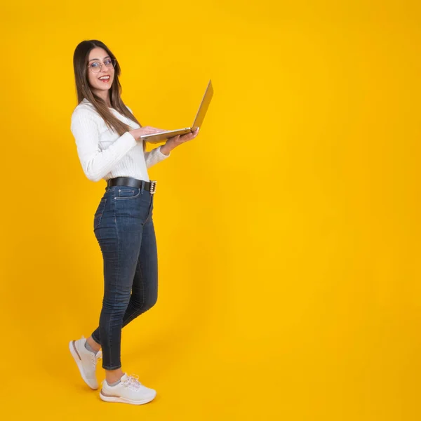 Full body length businesswoman holding laptop, standing over isolated yellow background. Social media post design concept, copy space. Smiling beautiful girl using notebook, computer.